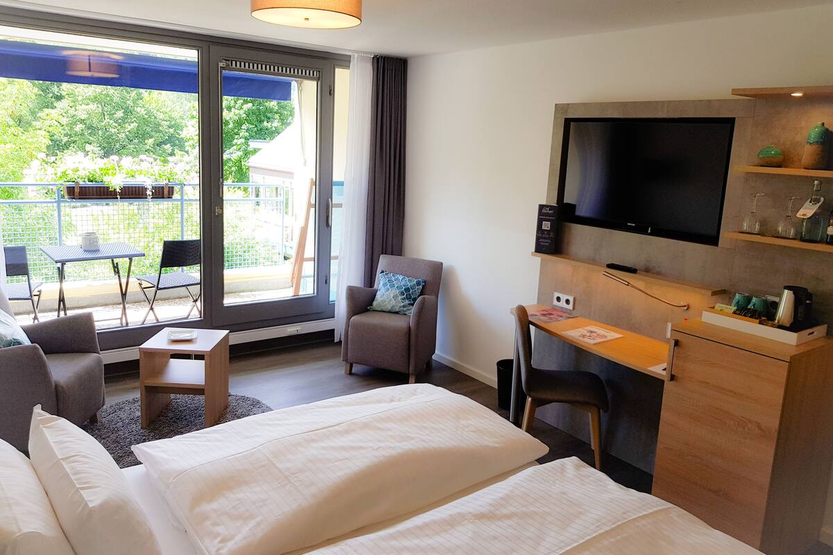 Habitación de hotel con cama, escritorio, silla, televisor y balcón con mesa y sillas.