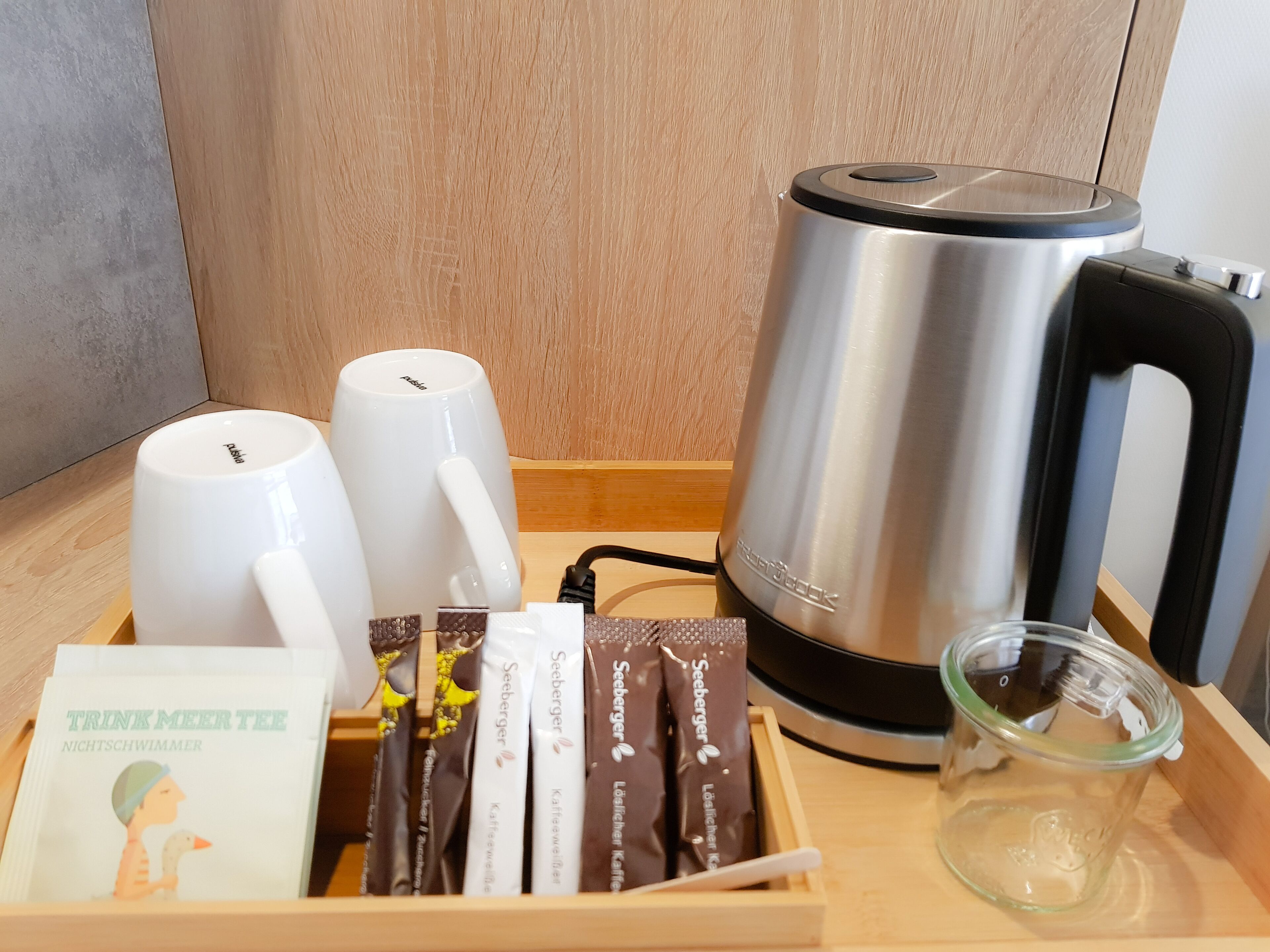 Una bandeja de madera con una cafetera, dos tazas blancas, un vaso y varios sobrecitos de Seeburger.