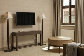A hotel room with a flat-screen TV mounted on the wall, a console table with a lamp, a round ottoman, and a view of the city through a window. - The Cloud One Dresden-Frauenkirche (Dresden)