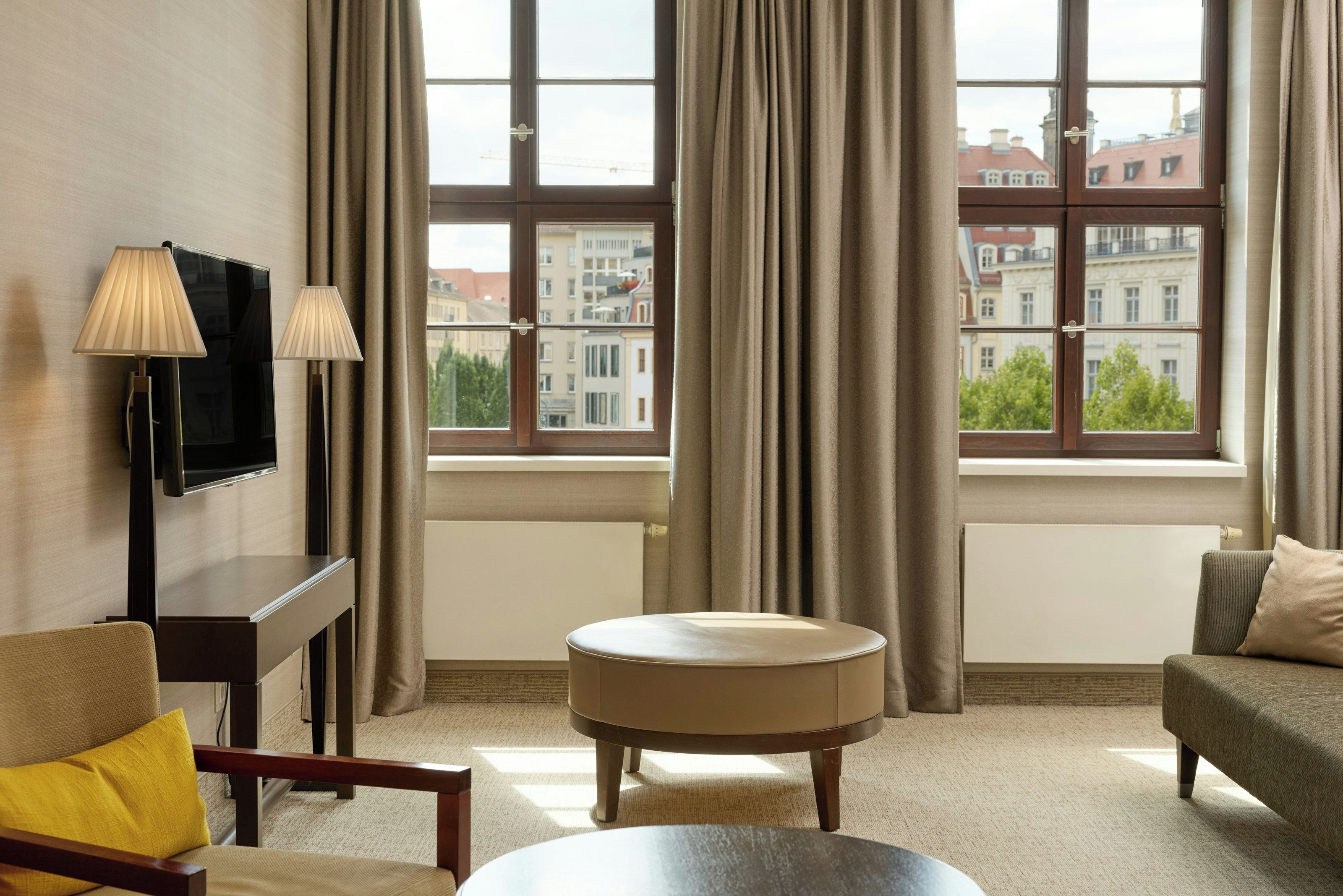 A hotel room with a large window, a flat-screen TV, two lamps, a round table, and a sofa with a yellow pillow.
