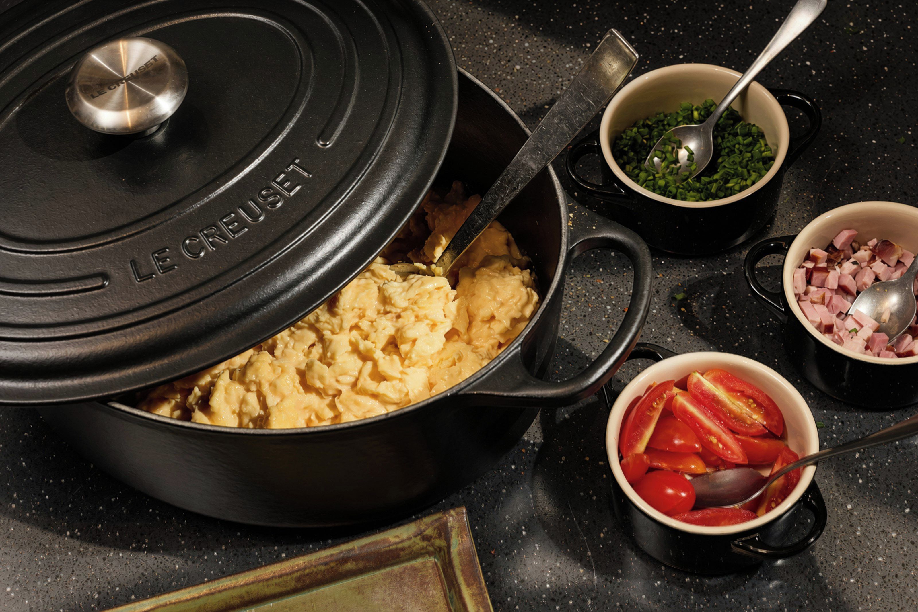 Una cocina con una olla grande en la estufa, un tazón de cebollines picados, un tazón de jamón en cubitos y un tazón de tomates en rodajas.