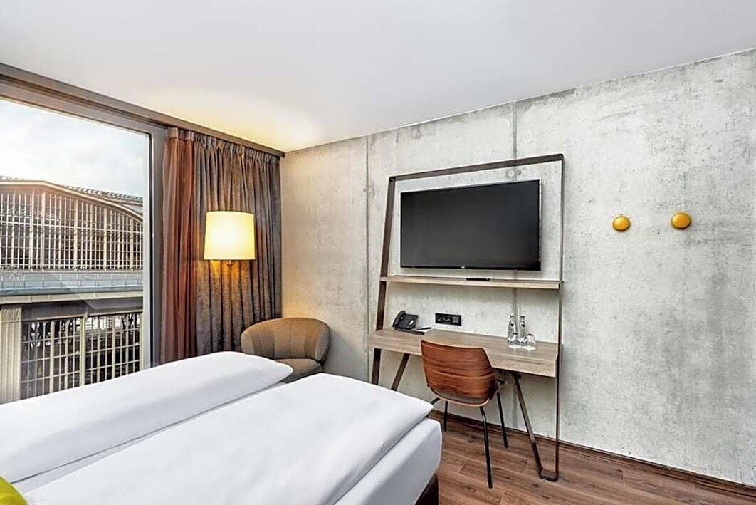 Ein Hotelzimmer mit Bett, Schreibtisch mit Stuhl, Fernseher und einem Fenster mit Vorhängen.
