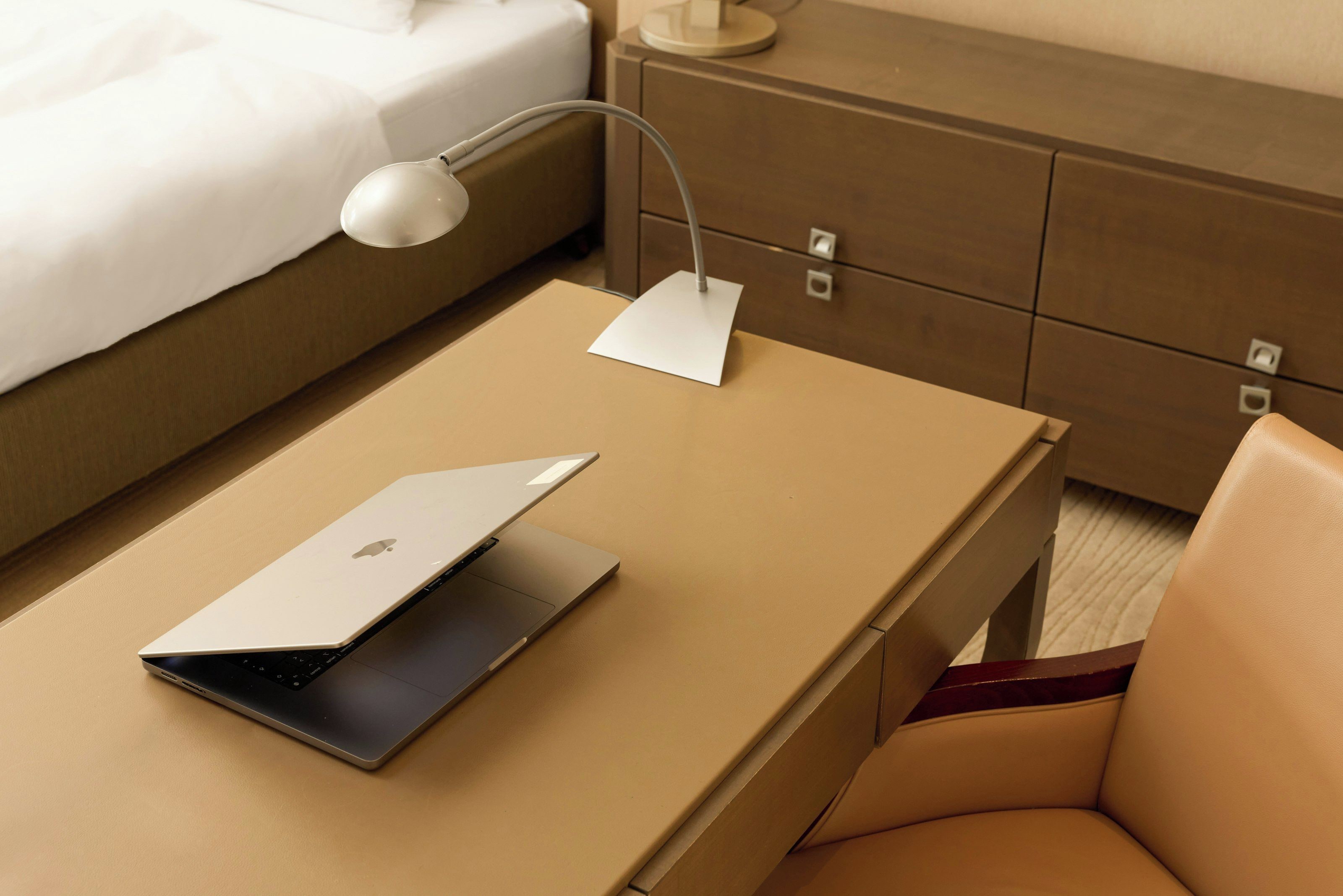 Een laptop op een beige tafel met een witte bureaulamp, een wit kussen op een bed en een houten nachtkastje met lades.