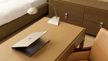 Een laptop op een beige tafel met een witte bureaulamp, een wit kussen op een bed en een houten nachtkastje met lades.
