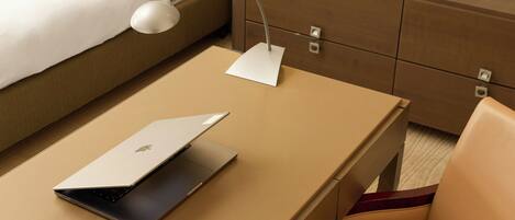 A laptop on a beige table with a white desk lamp, a white pillow on a bed, and a wooden nightstand with drawers.