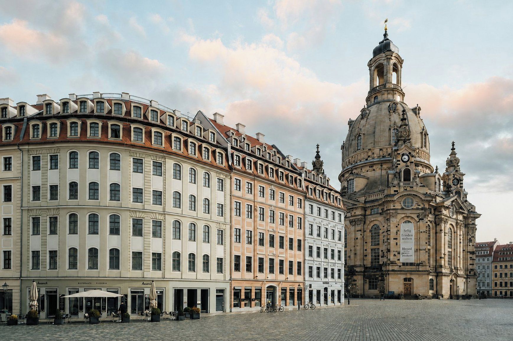 Ein historisches Gebäude mit einer markanten Kuppel und einem Uhrturm, umgeben von einem Kopfsteinpflasterplatz mit modernen Gebäuden.