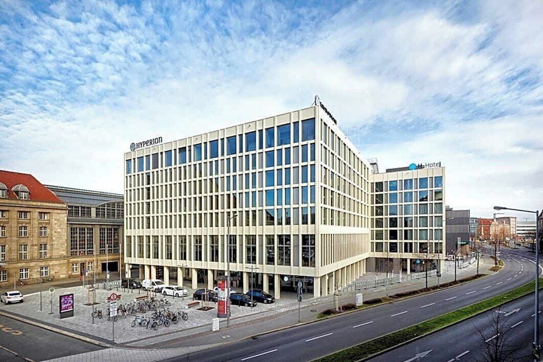 Ein modernes Gebäude mit Glasfassade, ein Schild mit der Aufschrift 'Hyperion Hotel' in der oberen rechten Ecke und ein Parkplatz mit Autos und Fahrrädern.