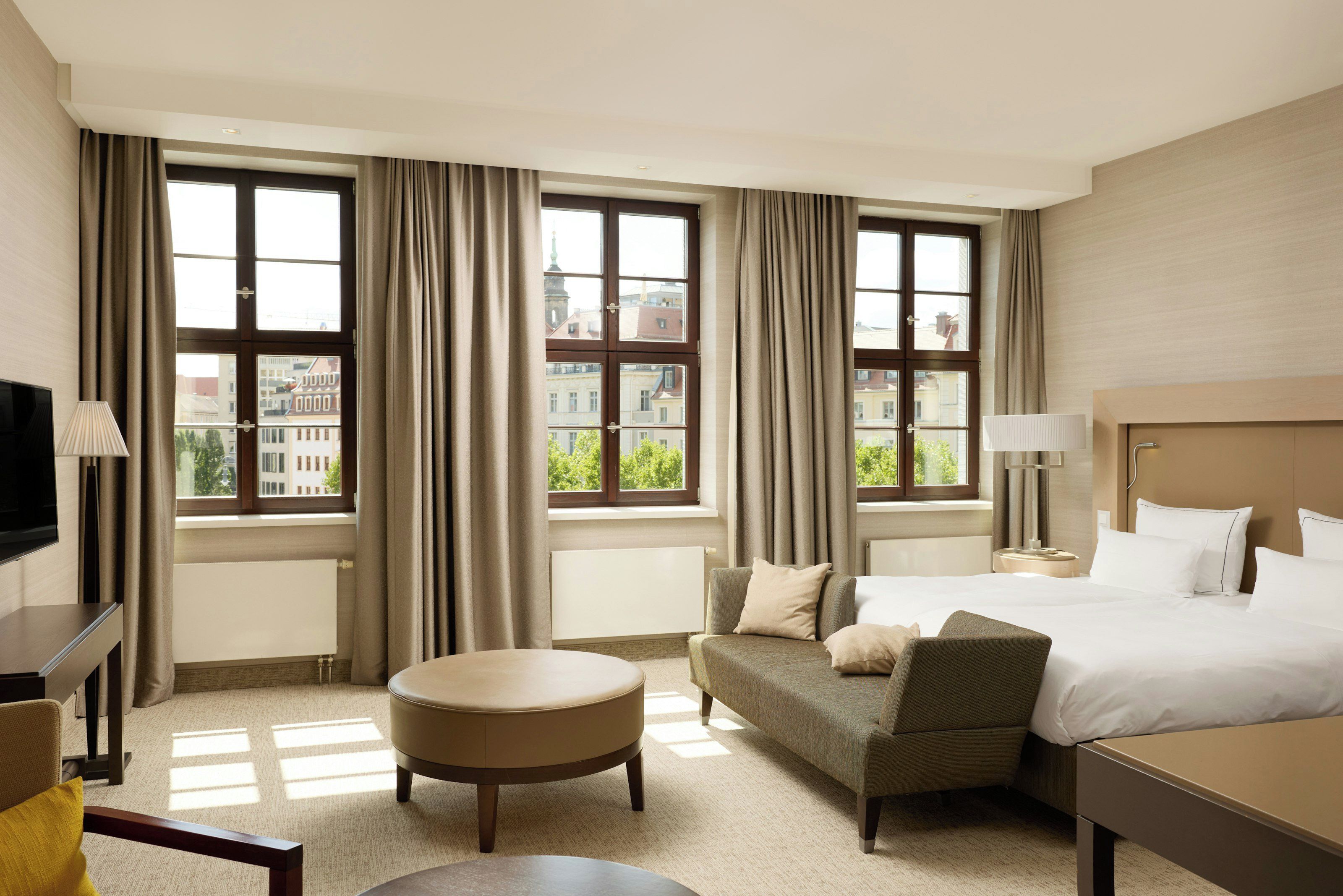 A hotel room with a bed, a sofa, a chair, a TV, and large windows with curtains.