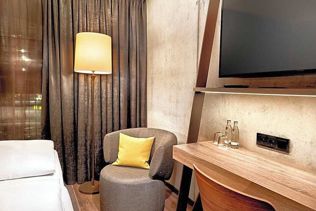A hotel room with a bed, a chair with a pillow, a table with bottles, and a TV mounted on the wall.