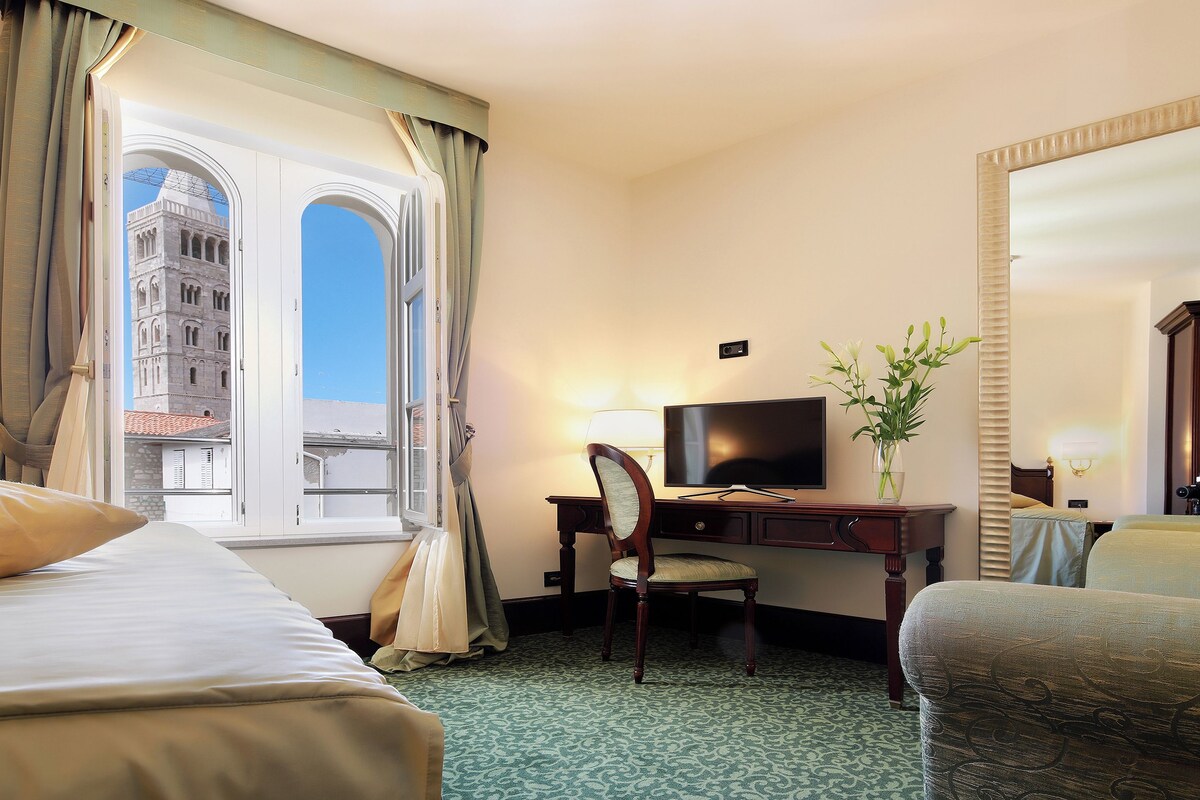 A hotel room with two beds, a desk with a TV, a chair, a vase with flowers, and a large mirror.