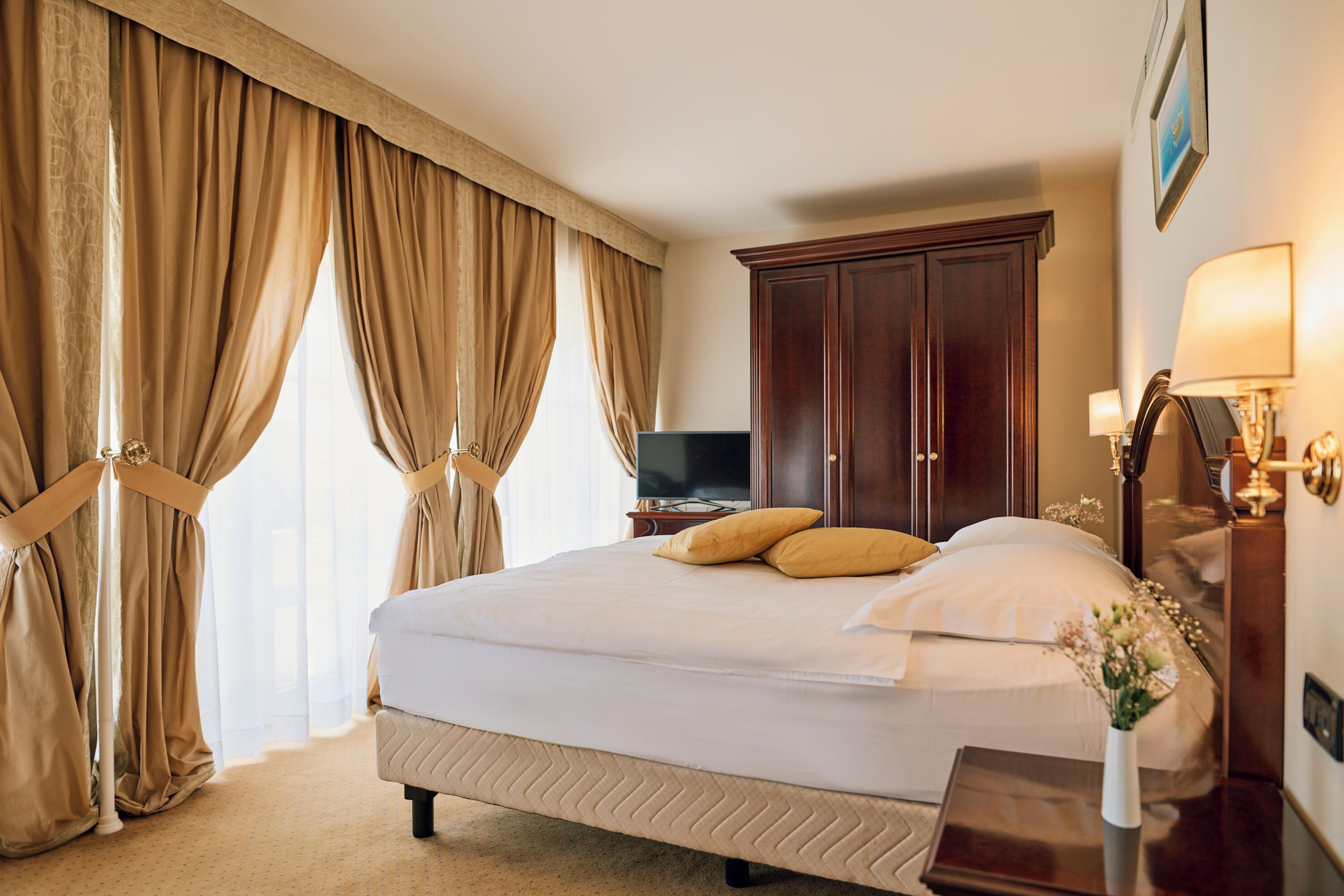 A hotel room with a large bed, beige curtains, a wooden wardrobe, a bedside table with a lamp, and a television.