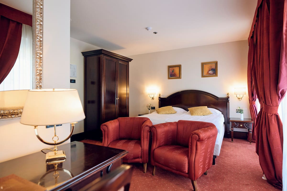 Une chambre d’hôtel avec un lit, deux fauteuils en cuir rouge, une table de chevet en bois, une lampe et un grand miroir.