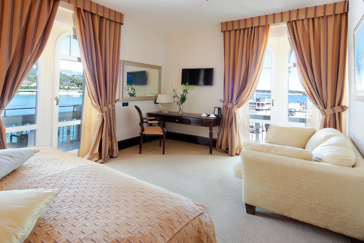 Une chambre d’hôtel avec deux lits, un bureau, une chaise et un canapé. Il y a une télévision et une fenêtre avec des rideaux.