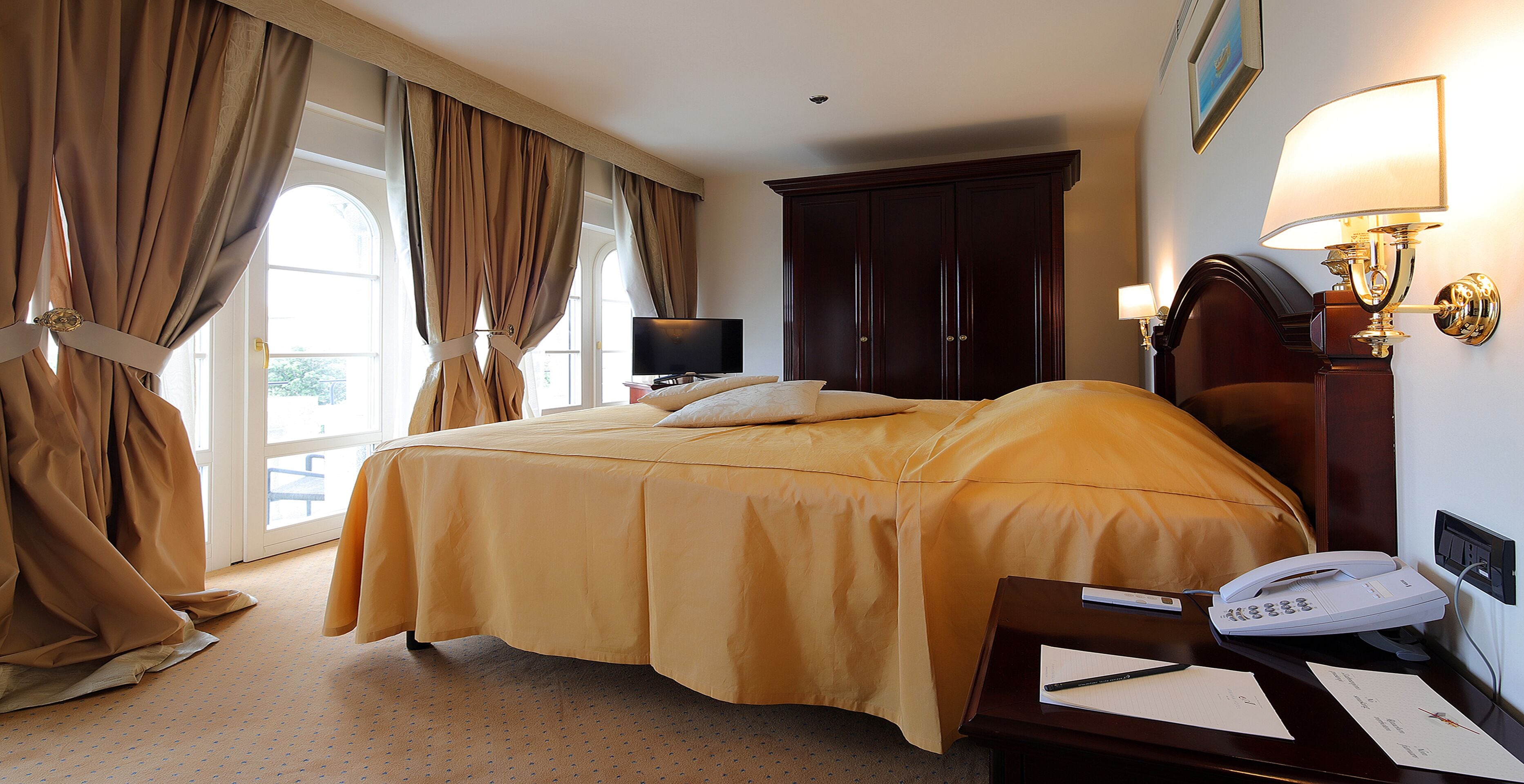 A hotel room with a bed, curtains, a TV, a desk with a phone, and a lamp.