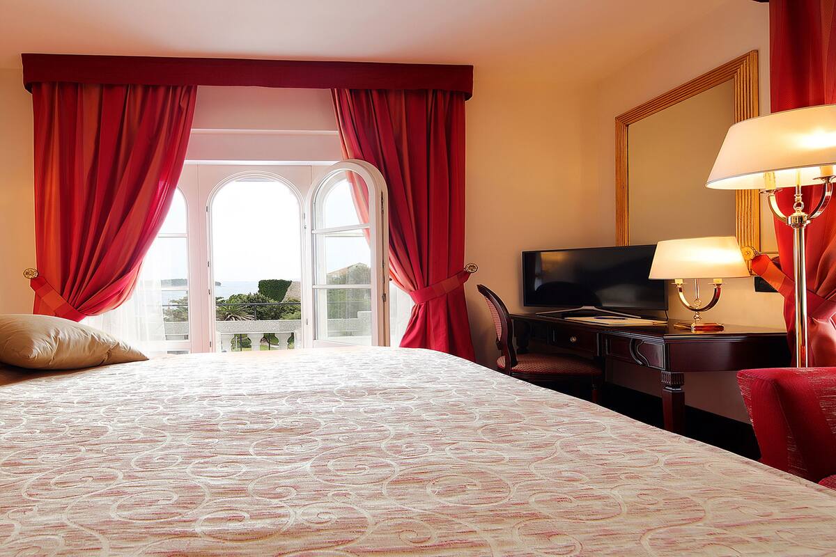 Une chambre d’hôtel avec un grand lit, un bureau avec une télévision et un balcon agrémenté de rideaux rouges.