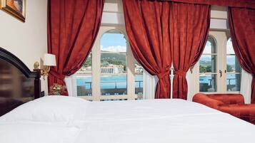 Habitación de hotel con una cama grande, cortinas rojas y vista a un cuerpo de agua y edificios.