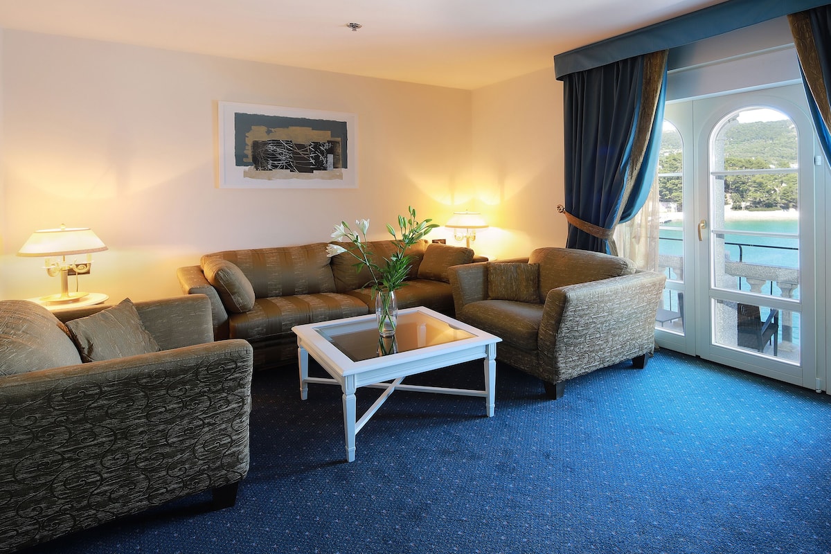 Une chambre d’hôtel comprenant un canapé, une table basse, deux lampes, un tableau encadré au mur et une vue sur l’extérieur grâce à une porte vitrée.
