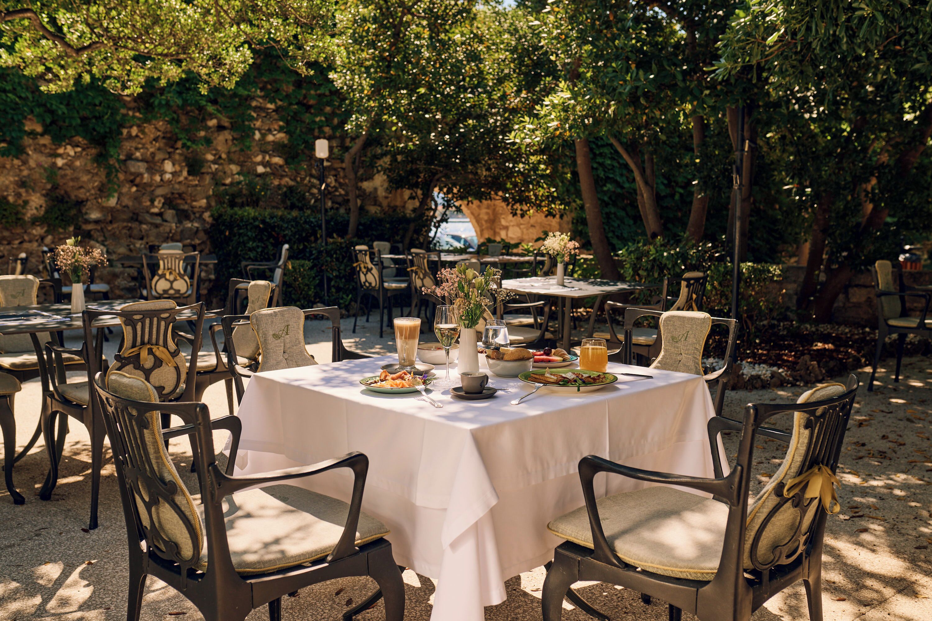 An outdoor dining area with round tables covered with white tablecloths, surrounded by chairs with cushioned seats.