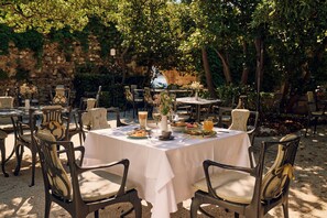 Un área de comedor al aire libre con mesas redondas cubiertas con manteles blancos, rodeada de sillas con asientos acolchados.