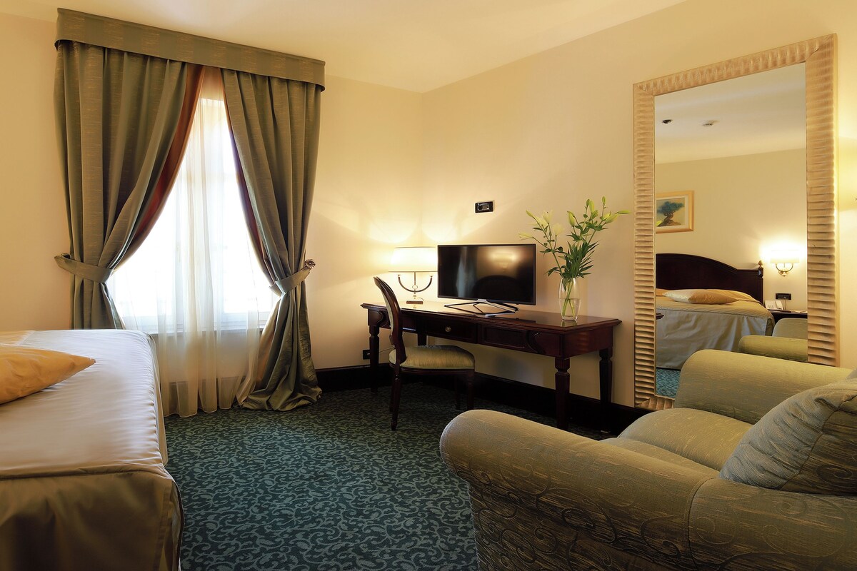 Habitación de hotel con cama, escritorio con lámpara, silla, espejo y ventana con cortinas.