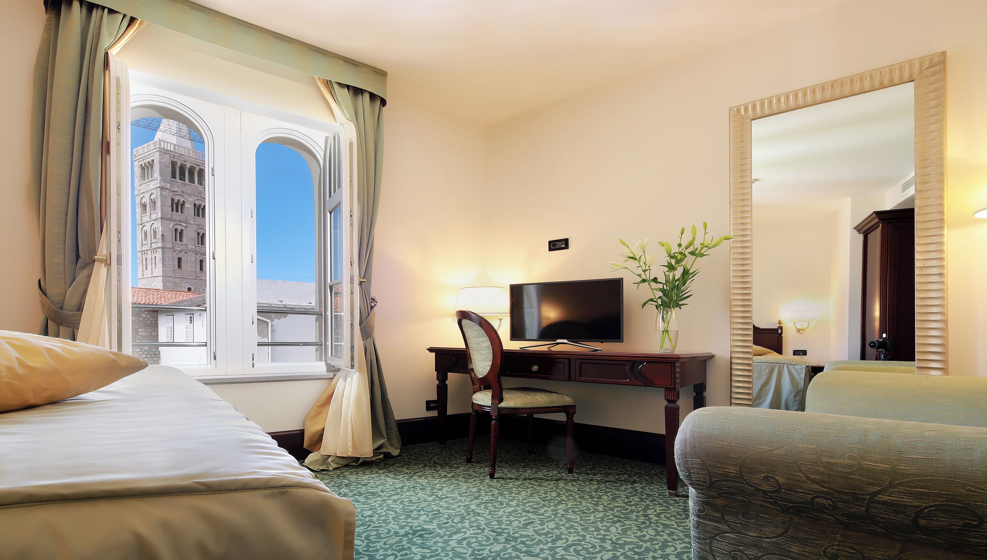 Une chambre d’hôtel avec deux lits, un bureau avec une télévision, une chaise, un vase de fleurs et un grand miroir.