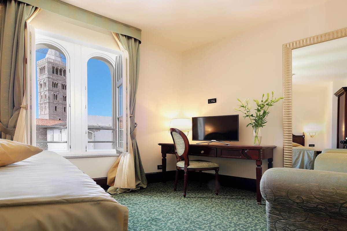 Une chambre d’hôtel avec deux lits, un bureau avec une télévision, une chaise, un vase de fleurs et un grand miroir.