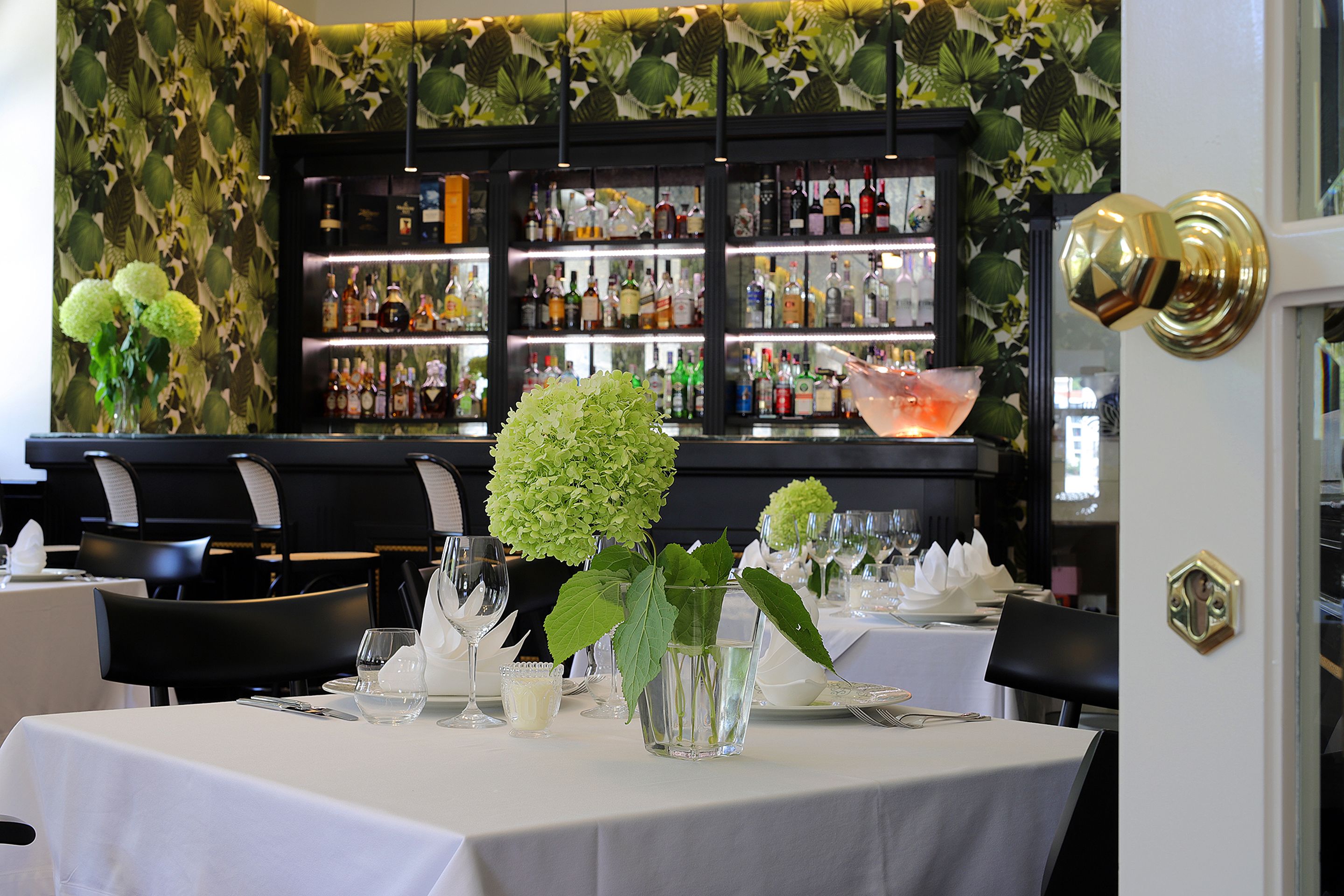 A restaurant interior with a bar area in the background. Tables are set with white tablecloths, and there are green plants on the tables.