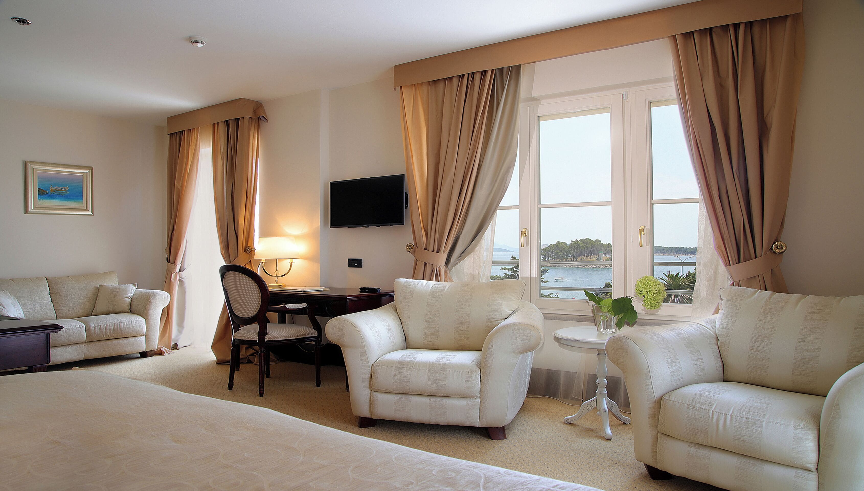 Une chambre d’hôtel comprenant un lit, deux fauteuils, un bureau avec une lampe, une télévision et une fenêtre avec des rideaux.