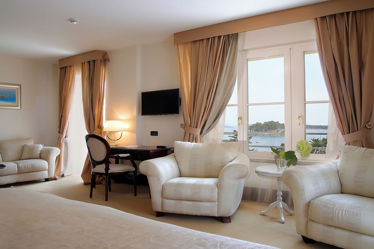 Une chambre d’hôtel comprenant un lit, deux fauteuils, un bureau avec une lampe, une télévision et une fenêtre avec des rideaux.