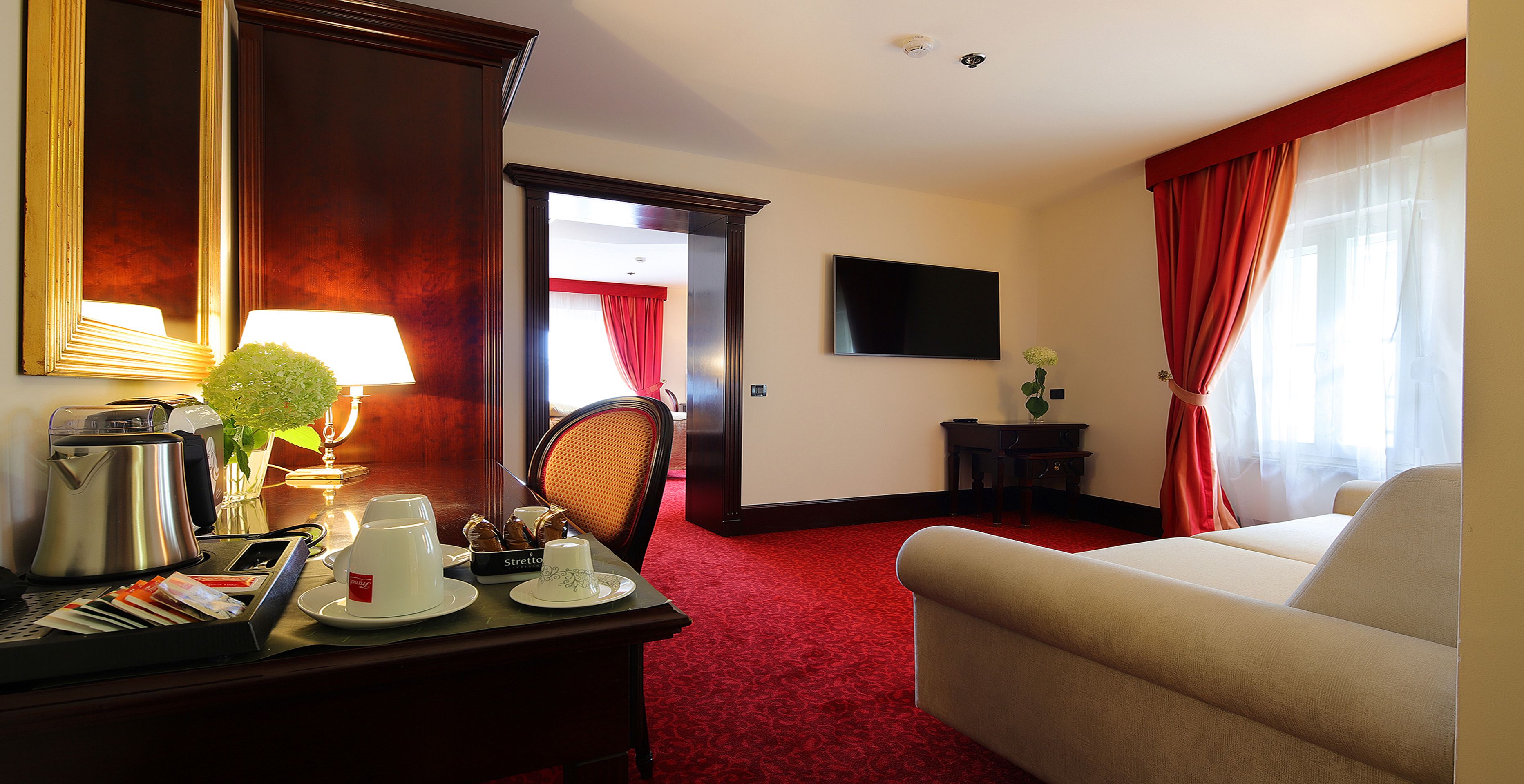 Een hotelkamer met een rood tapijt, een bank, een bureau met een koffiezetapparaat, een telefoon en een televisie aan de muur.