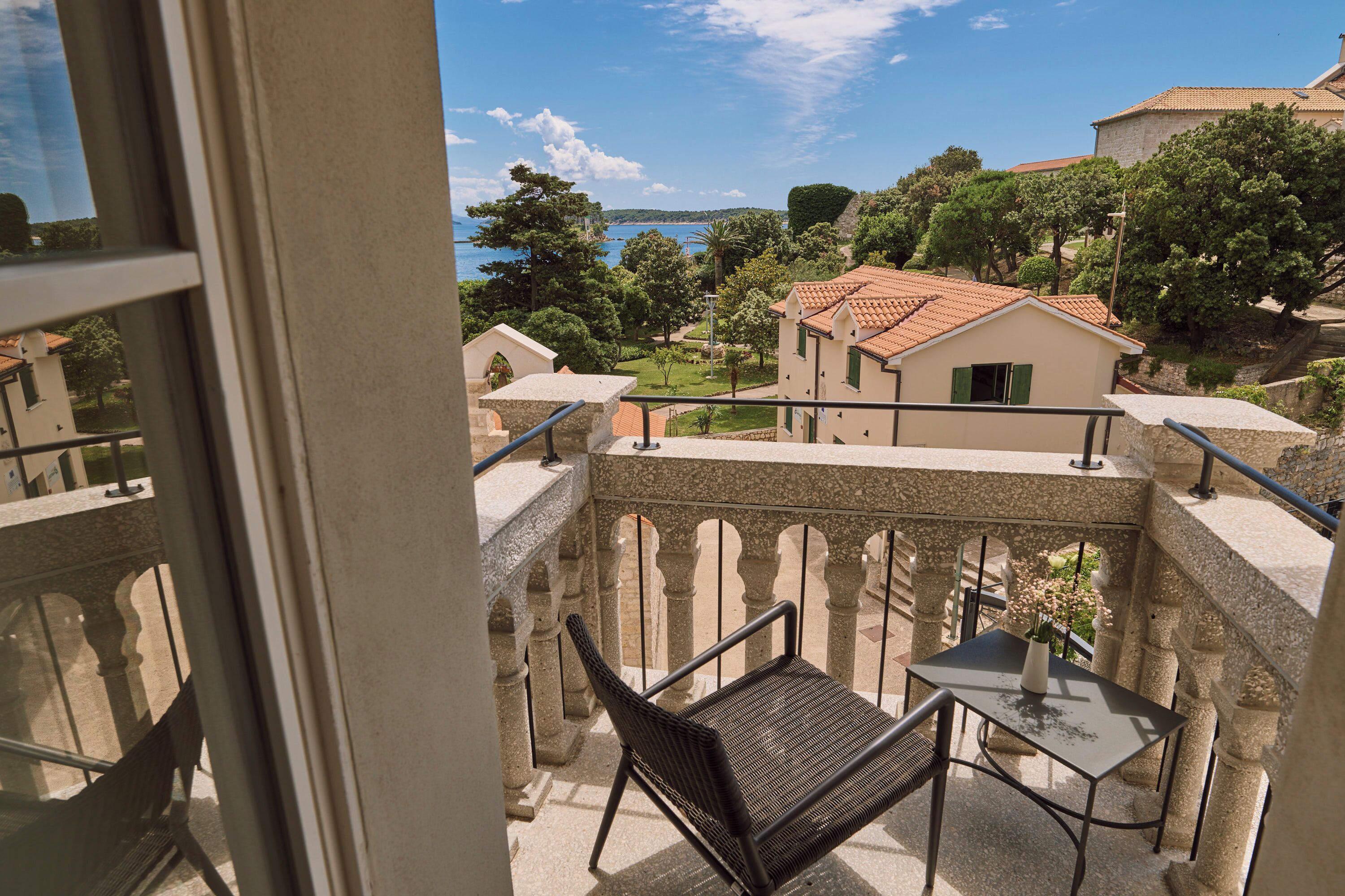 A balcony with a table and chair, overlooking a coastal area with buildings and trees.