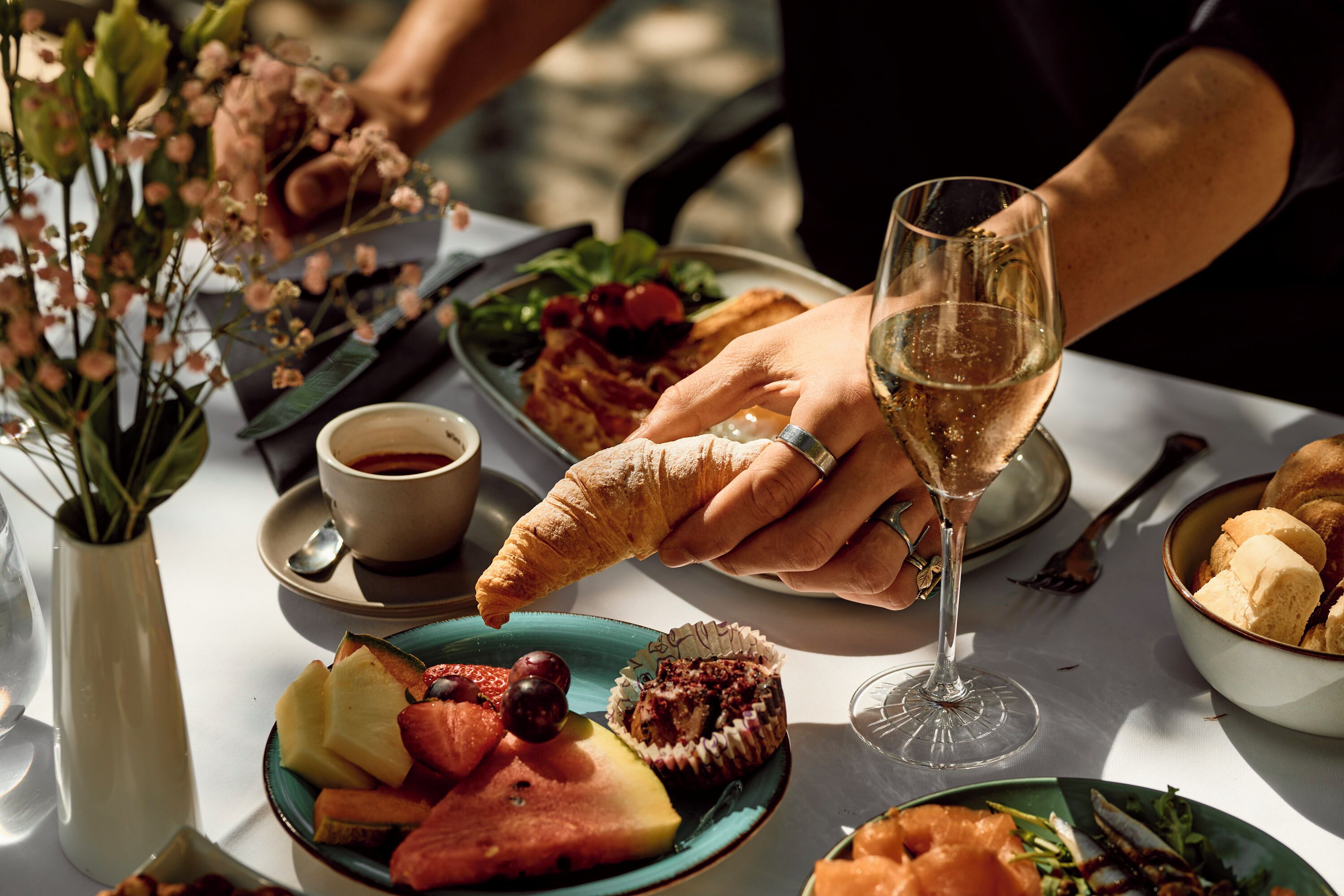 A dining table with various food items, a glass of wine, and a person holding a croissant.