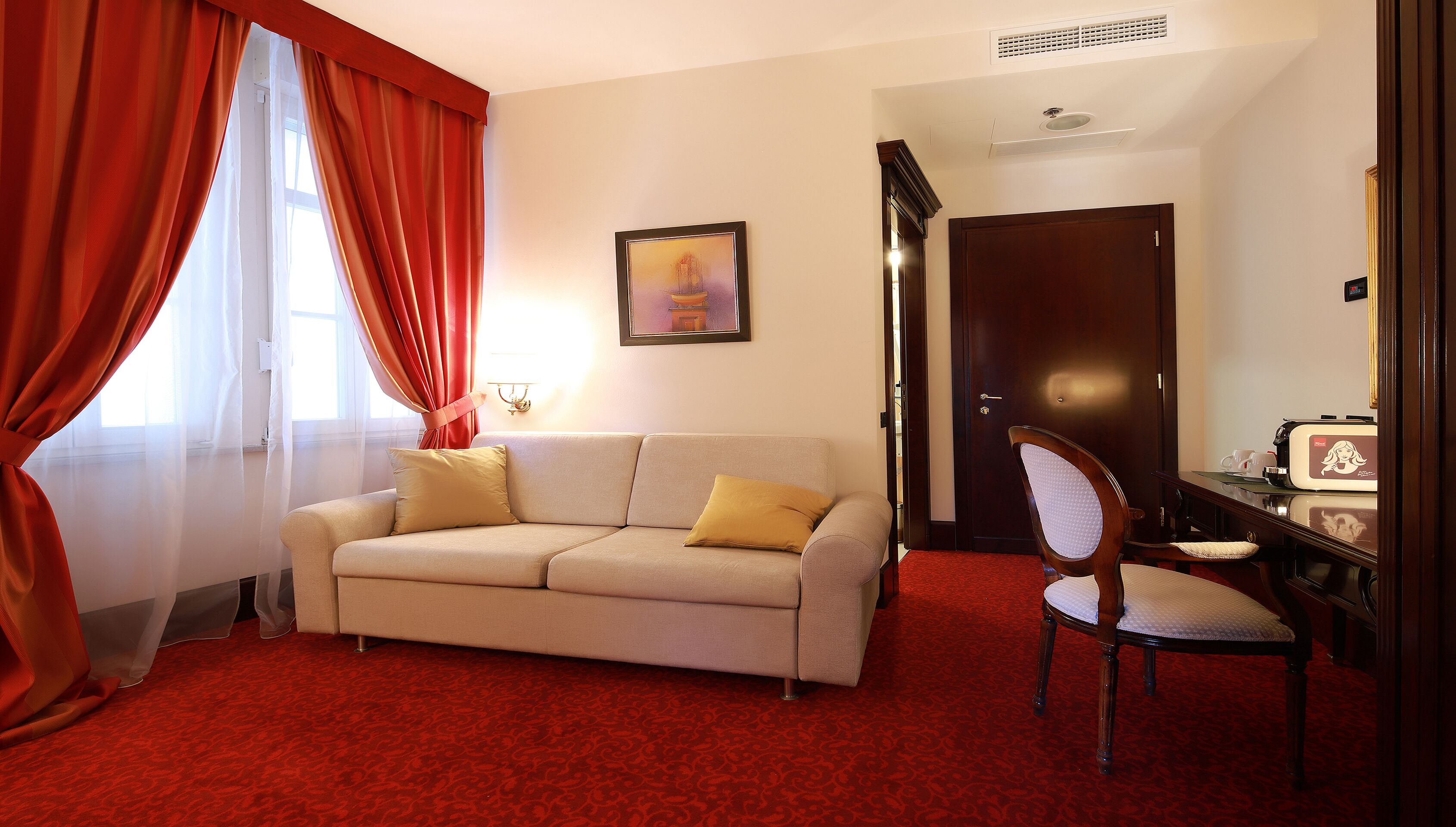 A room with a beige sofa, a wooden chair, a red carpet, a door, and a framed picture on the wall.
