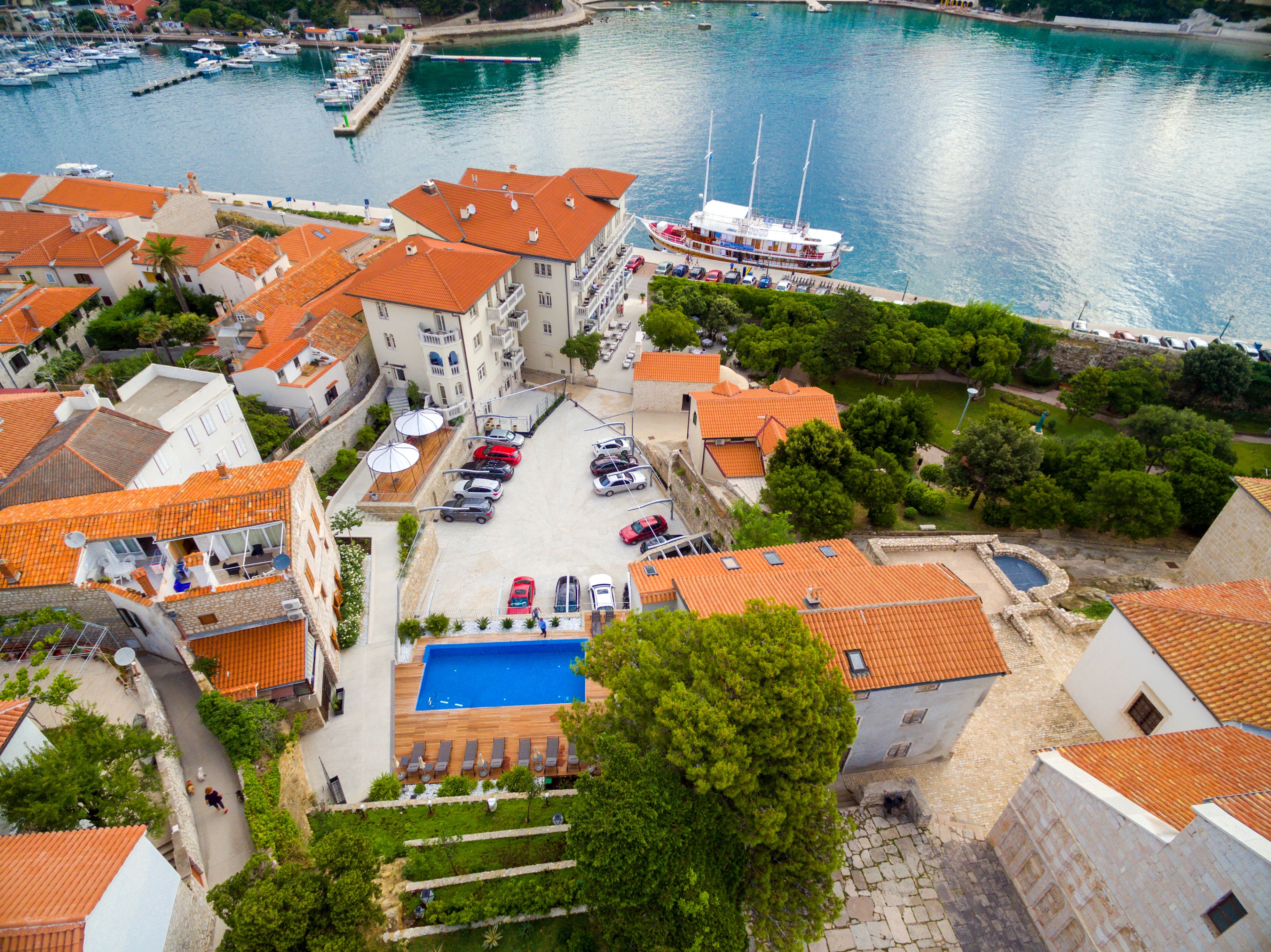 Una cittadina costiera con edifici a tetti in terracotta, un porto turistico con barche e una piscina con lettini.