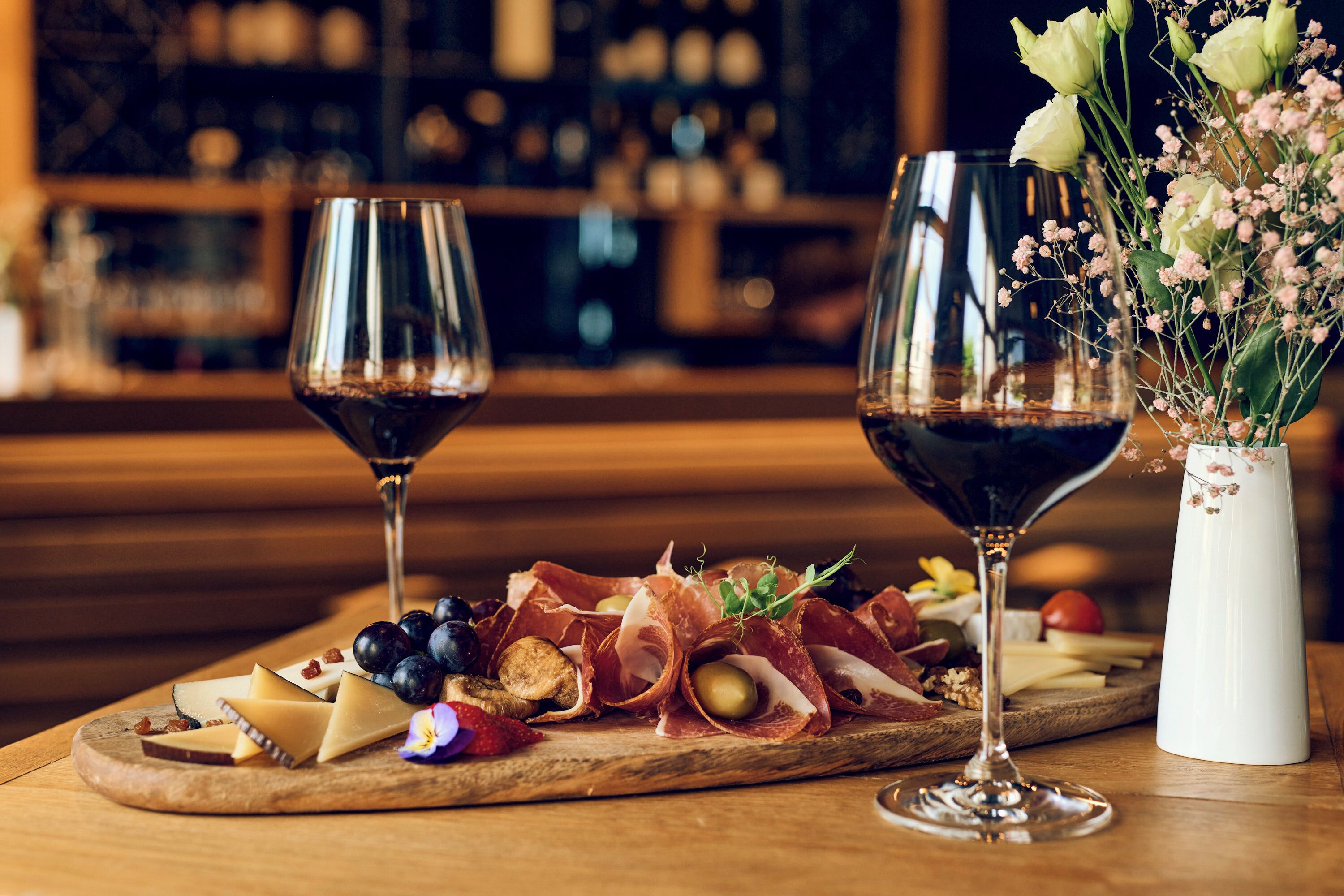 Ein Holzbord mit Käse, Trauben und Prosciutto, dazu zwei Weingläser und eine Vase mit Blumen.