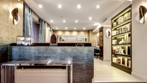 Un moderno vestíbulo de hotel con una recepción, un espejo grande y una estantería llena de libros y objetos decorativos.