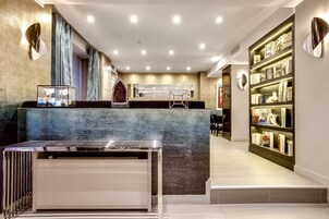 Un moderno vestíbulo de hotel con una recepción, un espejo grande y una estantería llena de libros y objetos decorativos.