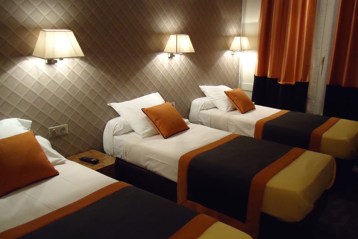 Ein Hotelzimmer mit drei Betten, jedes mit Kissen und Decke. An den Wänden Lampen, auf einem Nachttisch eine Fernbedienung.