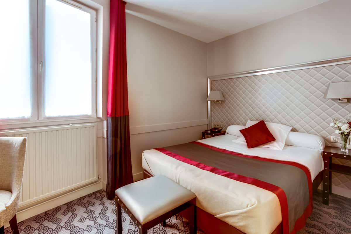 Ein Hotelzimmer mit einem Bett, einem Nachttisch mit Lampe, einem Sessel und einem Fenster mit Vorhängen.
