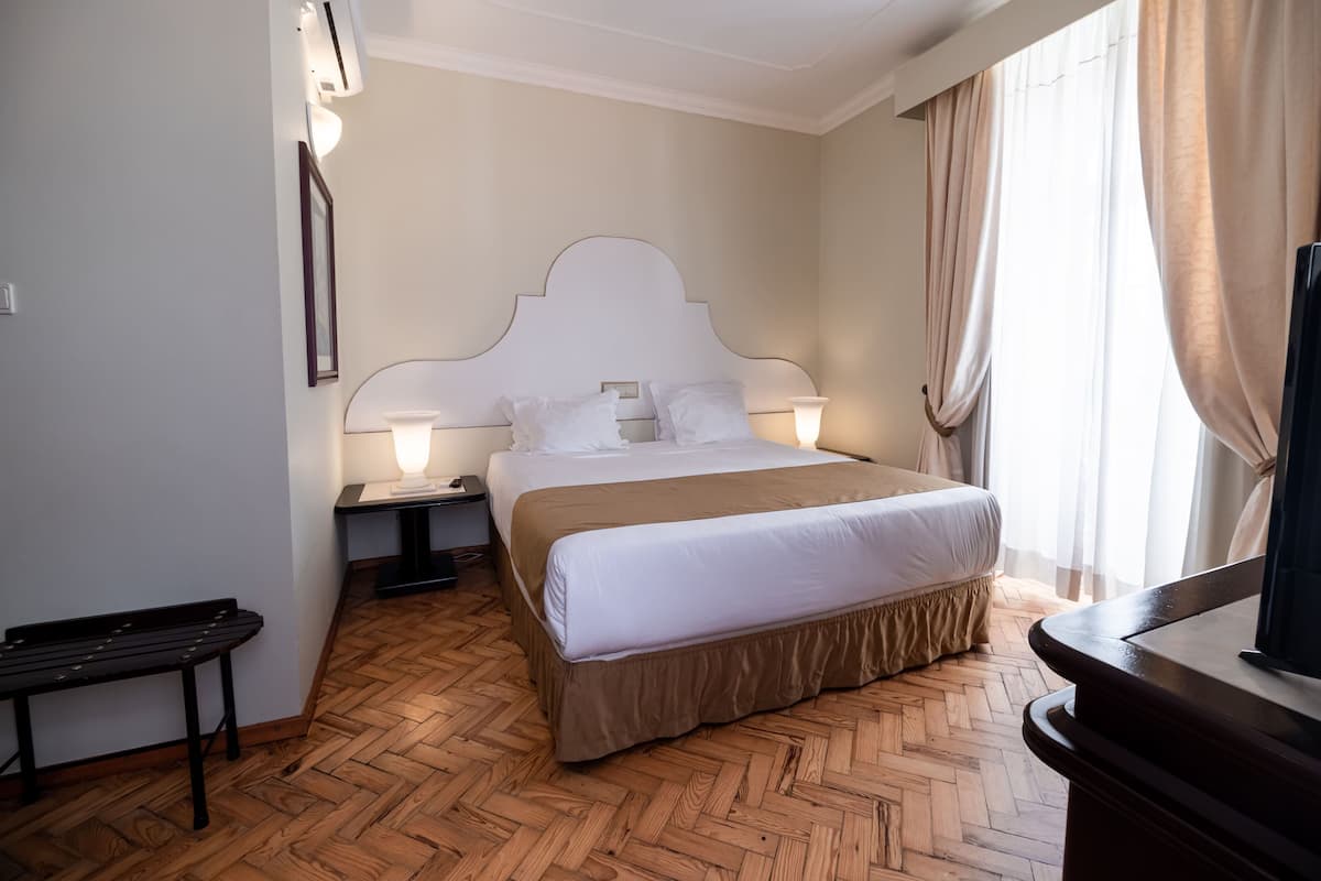 A hotel room with a large bed, two bedside lamps, a wooden floor, a bench, and a television.