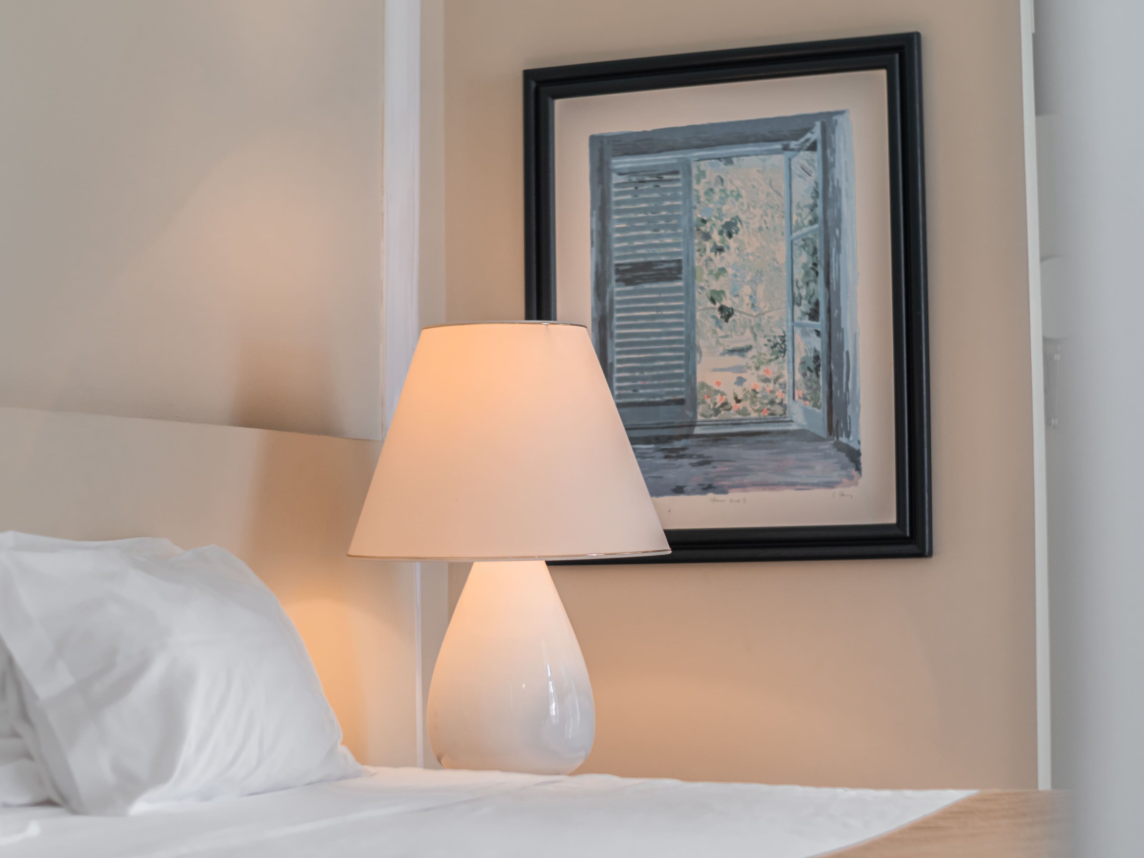 Cama bem arrumada com lençóis brancos, abajur branco e um quadro na parede retratando uma janela.