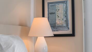 Cama bem arrumada com lençóis brancos, abajur branco e um quadro na parede retratando uma janela.