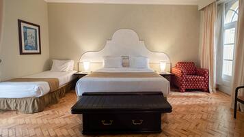 A hotel room with a large bed, two bedside tables, a red floral armchair, and a wooden bench.