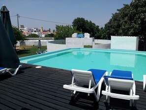 Ein rechteckiger Swimmingpool mit klarem, blauem Wasser, umgeben von einer Holzdielen-Terrasse. Auf der Terrasse stehen zwei weiße Liegestühle mit blauen Kissen.