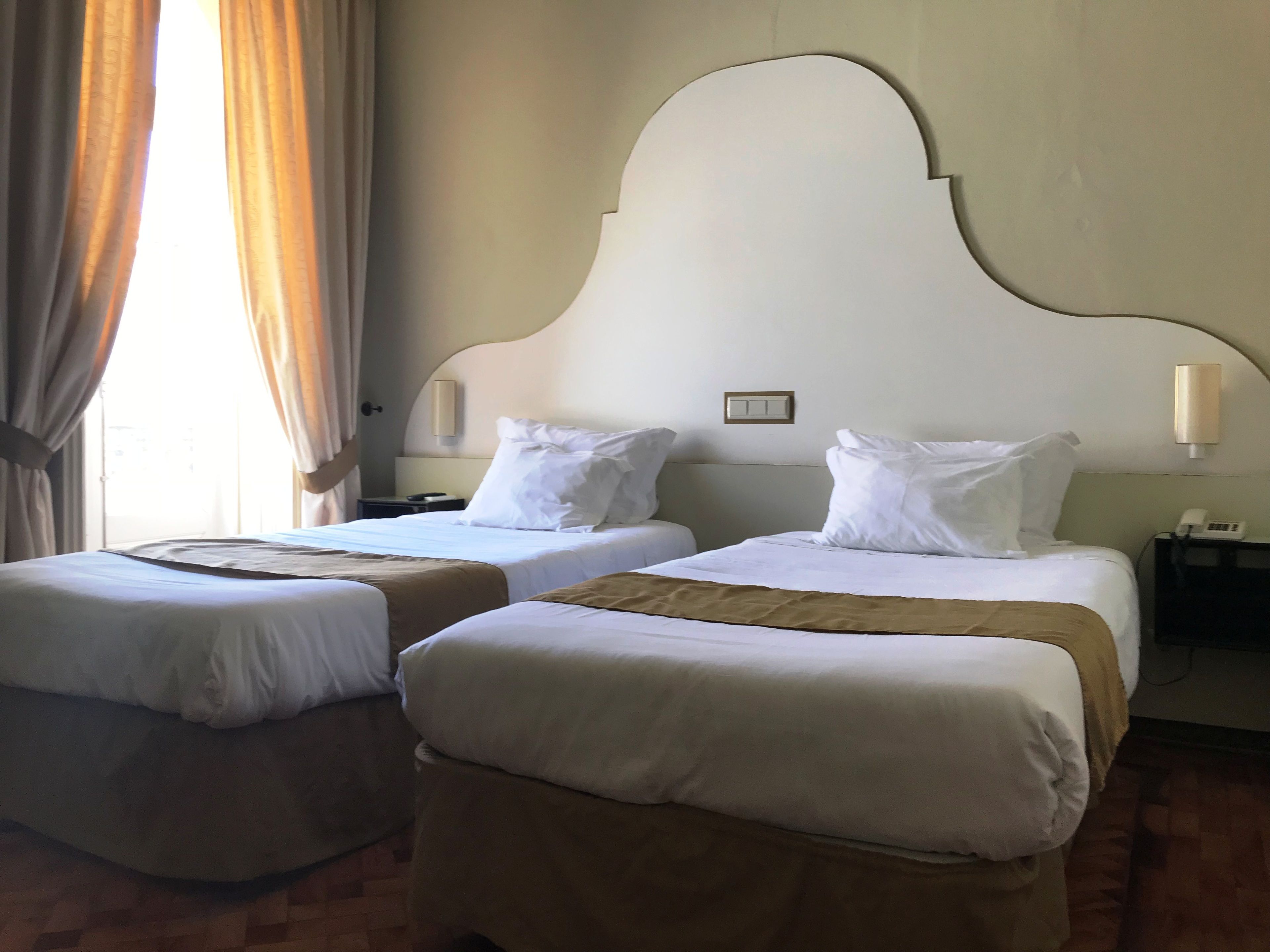 A hotel room with two beds, each with white linens and a beige throw blanket. There are two bedside tables with lamps, a wall-mounted light, and a phone on a nightstand.