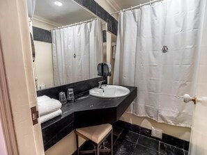 Ein Badezimmer mit einem weißen Waschbecken, einer schwarzen Arbeitsplatte und einem Spiegel darüber. Auf der Platte liegt ein weißes Handtuch und ein kleiner Hocker steht daneben.