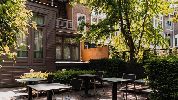 An outdoor patio with tables and chairs, surrounded by greenery and a building with a balcony. - Hotel Roemer (Amsterdam)