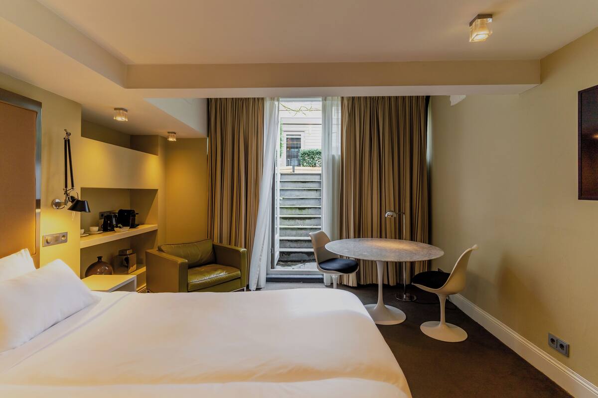 A hotel room with a large bed, a desk with a lamp, a chair, a small table, and a view of the outside through a window with curtains.