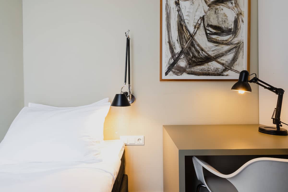 A neatly made bed with white linens, a black desk lamp, and a wall-mounted power outlet.