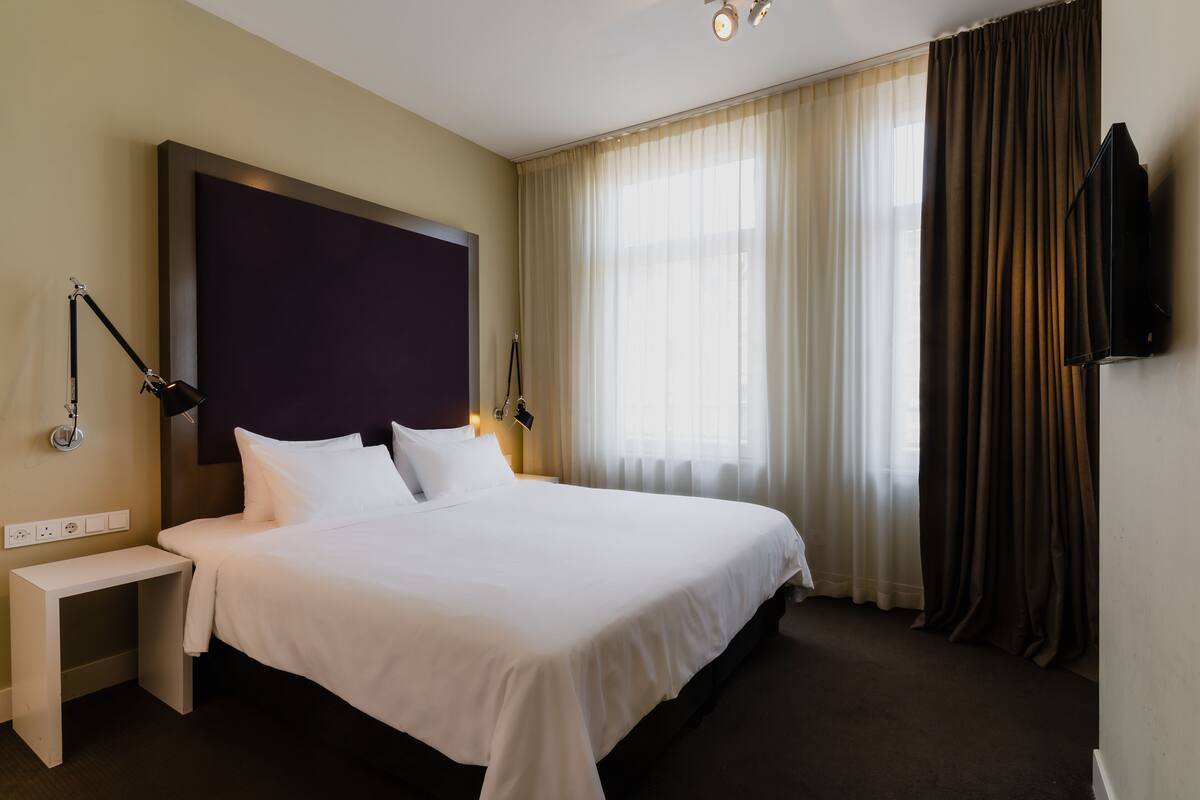 A hotel room with a large bed, bedside tables, a flat-screen TV, and a window with curtains.